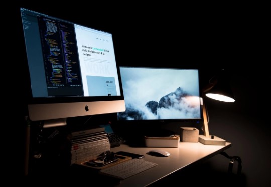 A creative technology workspace with code, a website interface, dual monitors, and a desk lamp